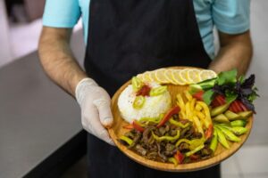 A chef holding a wooden platter with a colorful meal, including rice, grilled meat, and fresh vegetables.